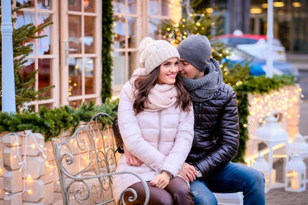 Happy romantic couple wearing warm clothes, enjoying spending time together on a date, hugging while sitting on a bench on a decorated street at Christmas timeの写真素材