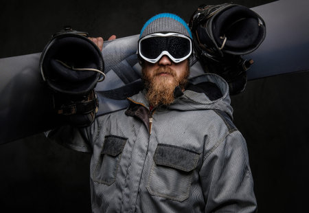 Brutal man with a red beard wearing a full equipment holding a snowboard on his shoulder, isolated on a dark textured background.の写真素材