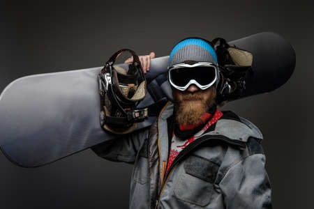A brutal man with a red beard wearing a full equipment holding a snowboard on his shoulder, isolated on a dark background.の写真素材