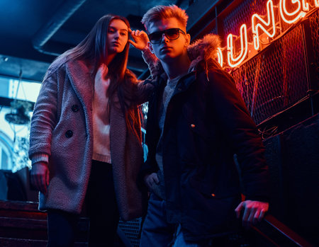 Stylish couple wearing warm clothes standing on the staircase to the underground nightclub, a...