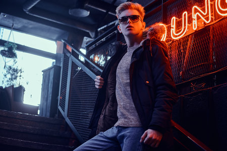 Young confident guy wearing coat with fur hood and sunglasses standing on the staircase to the...