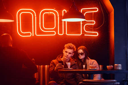 Trendy dressed young stylish couple sitting in a cafe with the industrial interior, chatting with friends or watching something on a phone