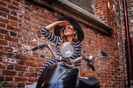 Cheerful woman wearing fashion clothing leaning on a black classic Italian scooter and looking away on an old street.の写真素材