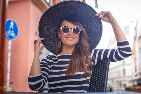Cheerful elegantly dressed young woman wearing a stylish hat and sunglasses sitting in a summer cafe.の写真素材