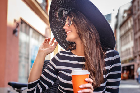 Cheerful fashionable woman wearing sunglasses and stylish hat holding a cup of coffee while sitting in a summer street cafe.の写真素材