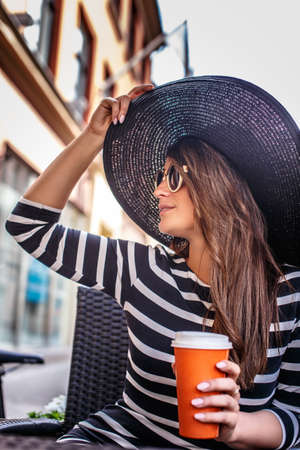Cheerful fashionable woman wearing sunglasses and stylish hat holding a cup of coffee while sitting in a summer street cafe.の写真素材