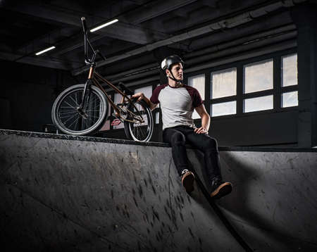 Young Bmx rider relaxing after practicing tricks with his bike in a skatepark indoorsの写真素材