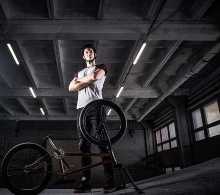 Full body portrait of a young man in protective helmet with his bike with his arms crossed standing in a skatepark indoorsの写真素材