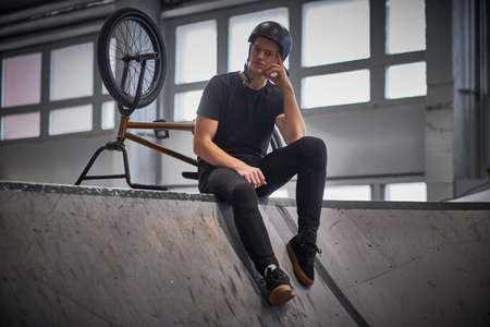 Pensive young man chilling after practicing tricks, sitting on a floor in a skatepark indoorsの写真素材