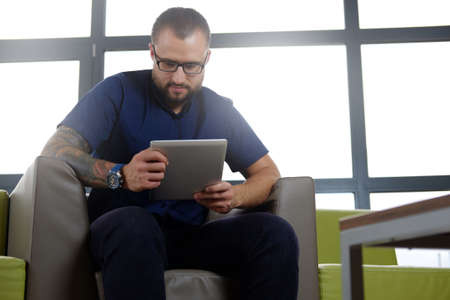 Bearded businessman dressed in a blue shirt sitting on the couch in the office, working with the...