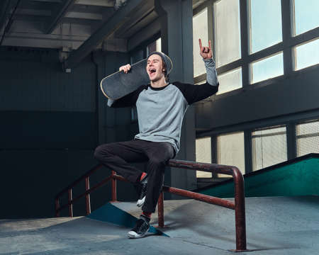 Cheerful young man wearing a shirt and hat holding his board, sitting on a grind rail in skatepark...