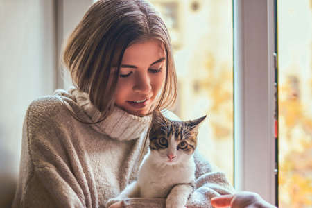 Beautiful girl in a warm sweater hugs her favorite cat sitting on the window sill next to the open...
