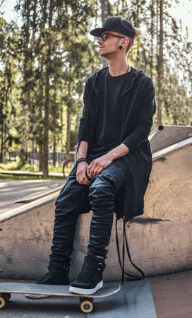 Young stylish skateboarder in a black hoodie and cap sits on the ramp in the skatepark in the summer...