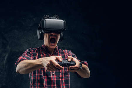 Young emotional man using VR headset and playing a video game with joystick in a studio against the background of the dark wallの写真素材