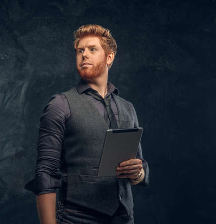 Handsome redhead man in formal wear holding a tablet in studio against a dark textured wall