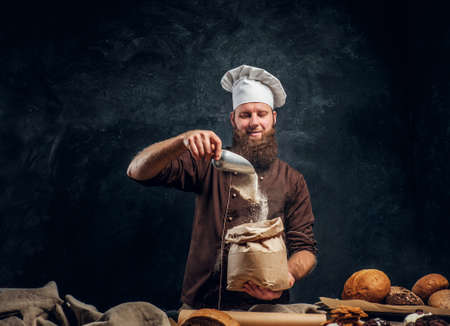 A bearded baker wearing a cook uniform sprinkling some flour in bag, standing next to a table, decorated with delicious bread loaves, baguettes and muffinsの写真素材