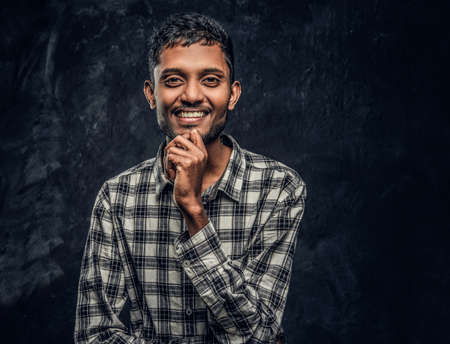 Portrait of a young Indian guy wearing a checkered shirt holding hand on chin, smiling and looking...