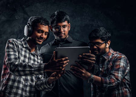 Three Indian friends watching something fun in a tablet standing in a dark studio against a black...