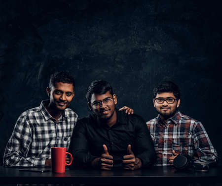 Three Indian student friends sitting together at the table, hugging and looking at a camera against...