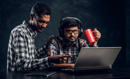 Two Indian student friends working together on a new project sitting at the table with a laptop against a dark textured wall.の写真素材