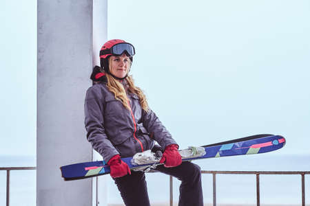 Portrait of a beautiful blonde woman in protective helmet and goggles holding skis while sitting on...