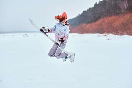 Cheerful woman wearing warming sportswear holding a snowboard and jumping on a snowy beachの写真素材