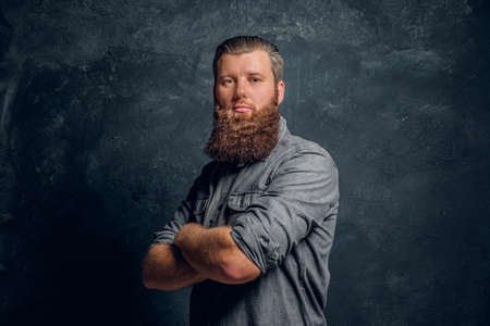 A man with a stylish haircut and beard wearing a gray shirt posing for a camera with his arms...