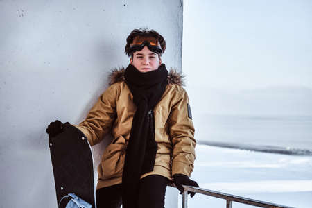 A teenage snowboarder dressed in warm clothes and protective goggles sitting on railing near the...