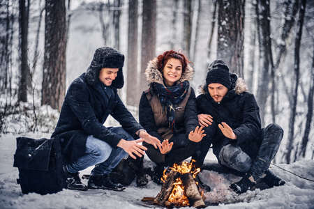 A group of happy friends staged a camping in the midst of a snowy forest, sitting around a campfire...