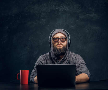 A hacker sits behind a laptop in a dark room and stares intently at the cameraの写真素材