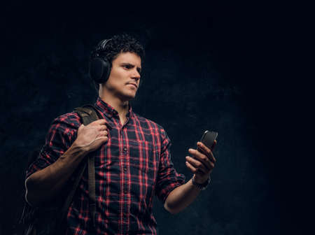 Pensive young guy in wireless headphones listens to music and keeps the smartphone in a studio against the background of the dark wallの写真素材