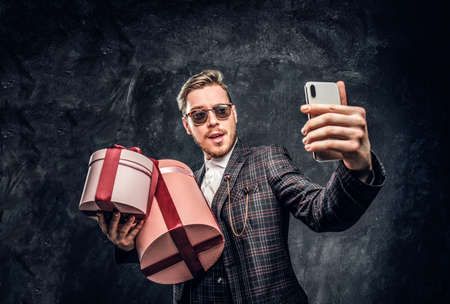 A young stylish man dressed in elegant suit holding gifts and taking selfie by smartphone in a dark...