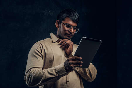 Pensive Indian male in eyewear and shirt using a tablet computer in studio against the background of the dark wallの写真素材