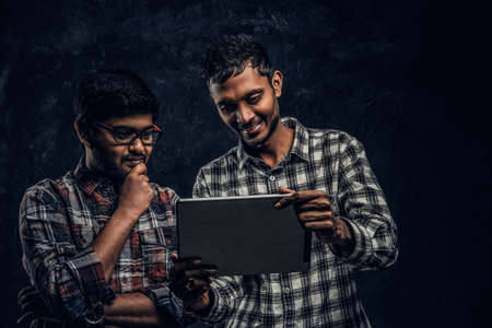 Three happy Indian friends in casual clothes stand in a hug and taking selfie on smartphone. Studio...