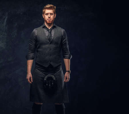 Handsome redhead man dressed in an elegant vest with tie and kilt in studio against a dark textured...