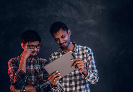 Three happy Indian friends in casual clothes stand in a hug and taking selfie on smartphone. Studio...