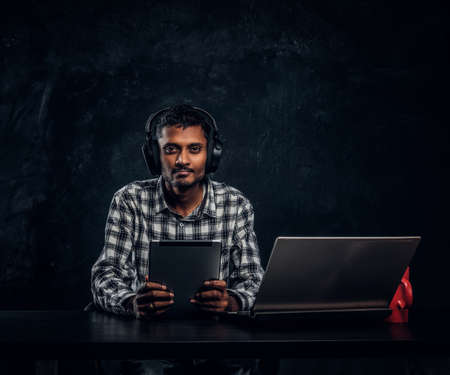 Indian student in headphones holding a tablet, sitting at a desk and looking at a camera. Studio photo against a dark textured wallの写真素材