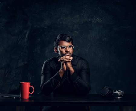 Indian student sitting at a desk with laptop, leaning chin on his hands and looking sideways. Studio photo against a dark textured wallの写真素材