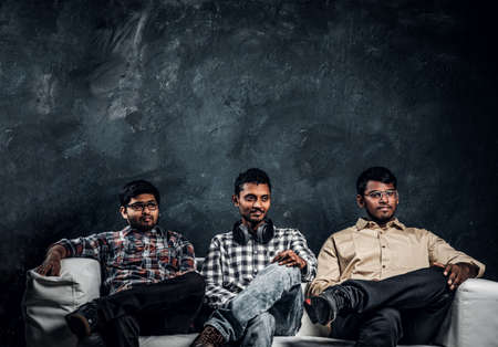 Three Indian student friends wearing casual clothes sitting on a couch against the dark wall