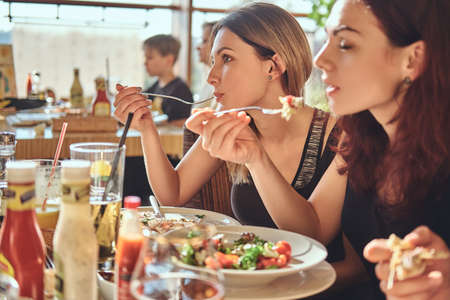 Two young girls eat salads sitting with their friends at the cafe during lunch breakの写真素材