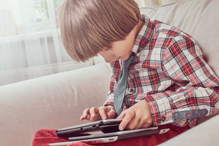 Young Caucasian schoolboy wearing a checkered shirt with tie using a digital tablet sitting on a couch at homeの写真素材