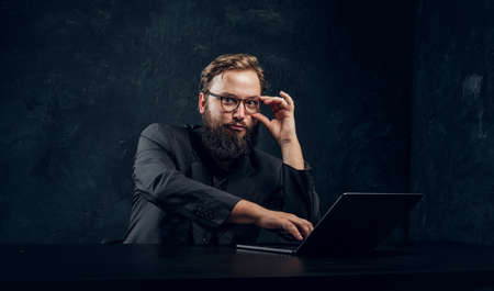 Bearded manager working behind a laptop and a cunning look at the camera holding glasses in the...