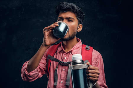 Young stylish hiker with backpack drinking a tea from a thermos. Studio photo against a dark textured wallの写真素材