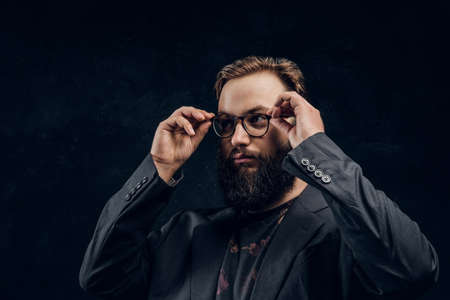 Portrait of a smart bearded guy wearing a black jacket corrects his glasses while standing in a dark...