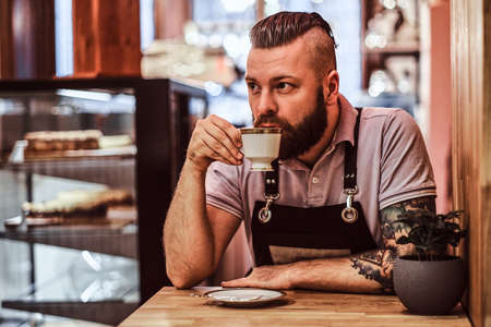 Handsome barista in apron drinking coffee during lunch break sitting at a table in the coffee shopの写真素材