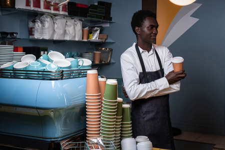African barista holding a cup with coffee while standing behind a counter in a coffee shop and looking sidewaysの写真素材