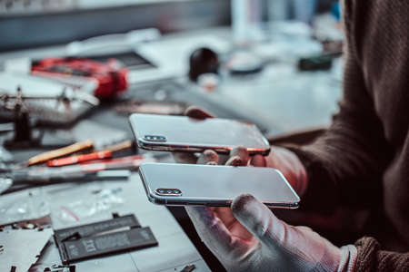 Electronic technician holds two identical smartphones for comparison, in one hand broken and in...