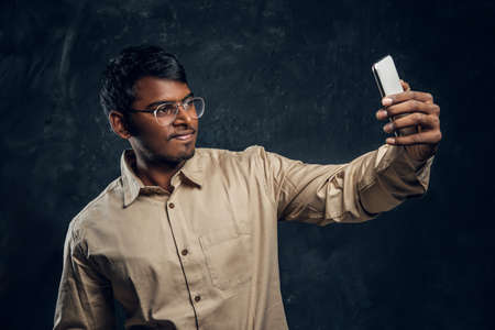 Handsome hindu student smiles and makes selfie on phone in studio on black backgroundの写真素材