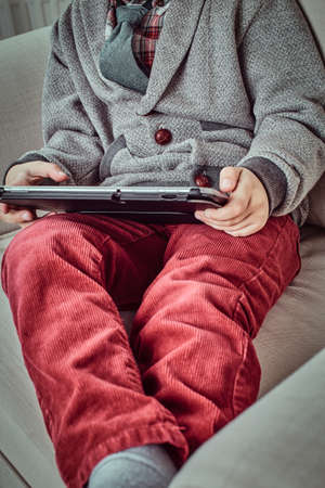 Young Caucasian schoolboy wearing elegant clothes using a digital tablet while sitting on a couch at home. Cropped imageの写真素材