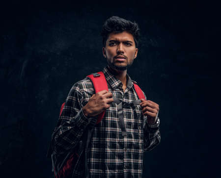 Portrait of a handsome Indian student with a backpack wearing a plaid shirt. Studio photo against a dark textured wallの写真素材